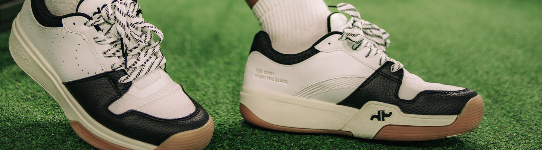 White and black low-top court shoes with patterned laces and gum soles, worn on a green outdoor pickleball court.