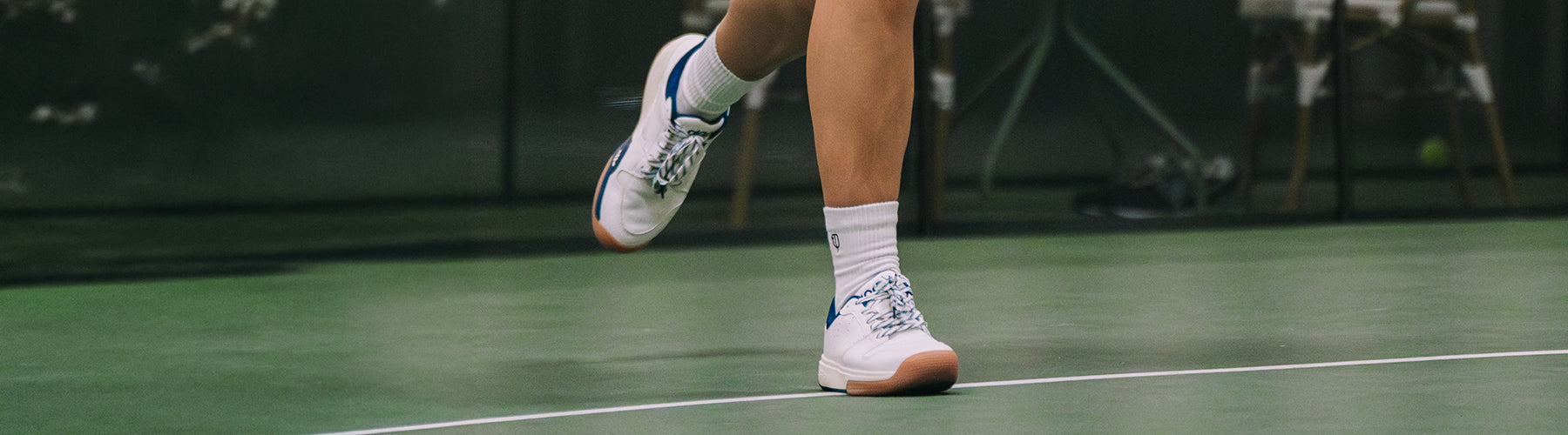 A pickleball player's feet mid-stride on an indoor hard court, wearing white and blue court shoes built for lateral movement and grip