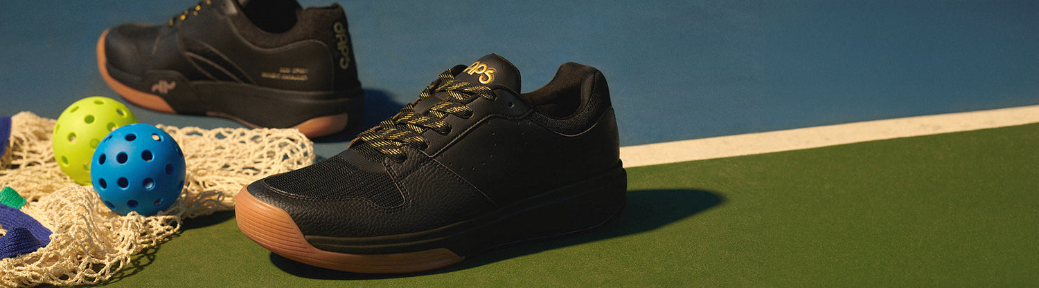 DAPS Desi Low men's pickleball shoes in black and gold on an outdoor hard court, surrounded by pickleballs and a net.