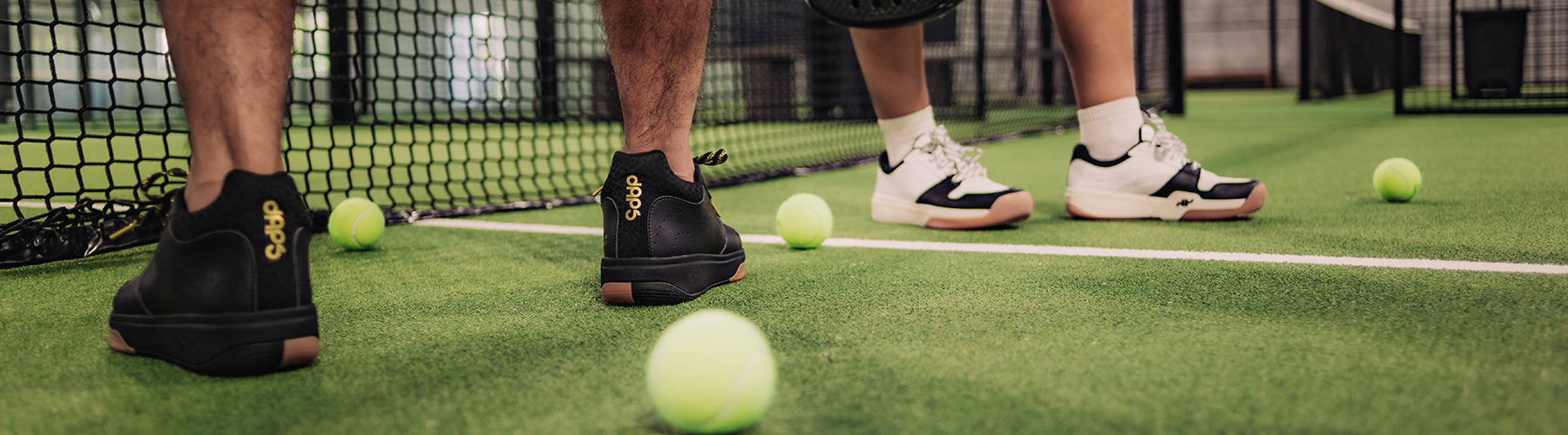 Two players in black and white DAPS pickleball shoes standing near the net, with pickleballs scattered across the court.