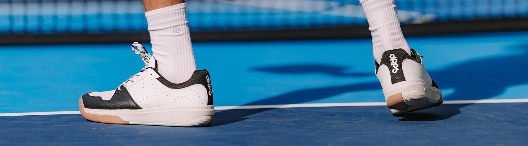 White and black DAPS court shoes worn on a blue pickleball court, with tall socks providing extra ankle coverage.