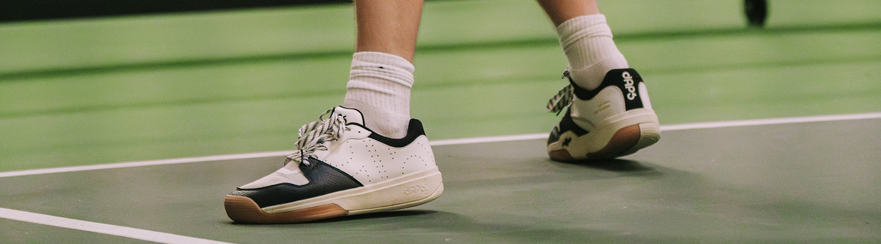 Close-up of a woman's feet wearing white and black pickleball court shoes on an indoor green court surface