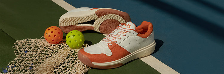 DAPS Desi Low pickleball shoes in burnt orange and white, styled on a pickleball court alongside wiffle balls and a net, showing why purpose-built court shoes outperform running shoes for pickleball.