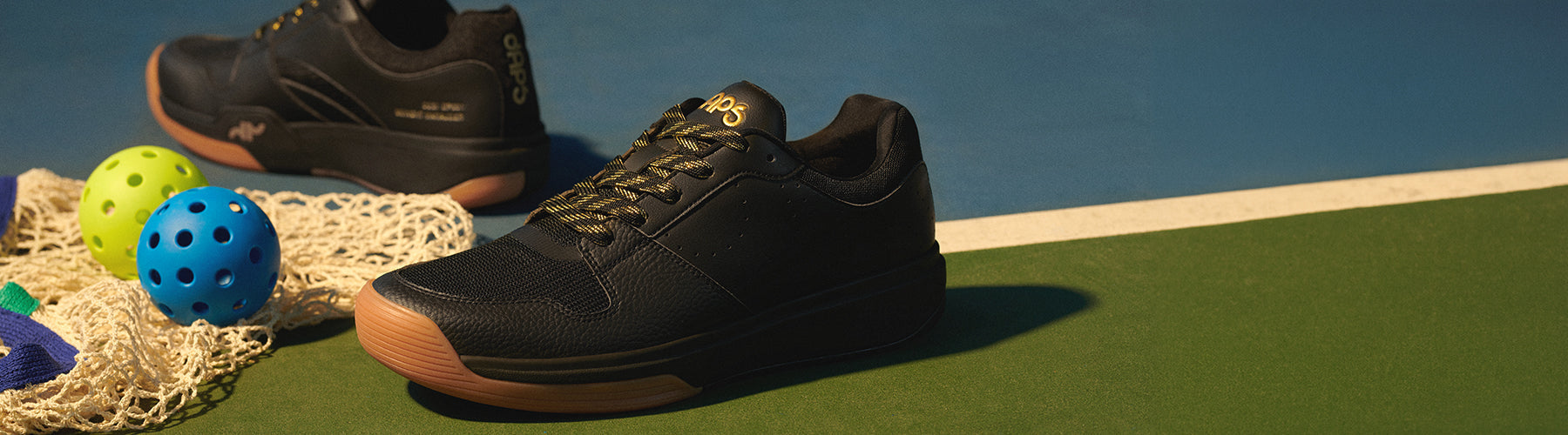 DAPS Desi Low men's pickleball shoes in black and gold on an outdoor hard court, surrounded by pickleballs and a net.