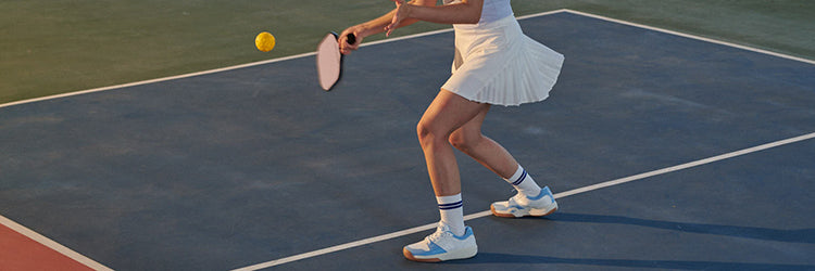 A pickleball player in a white pleated skirt and blue athletic shoes prepares to hit a forehand shot on an outdoor court