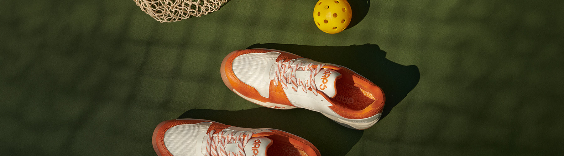 A pair of DAPS Desi Low outdoor pickleball shoes built for court traction, shown on a green outdoor court surface with a pickleball nearby