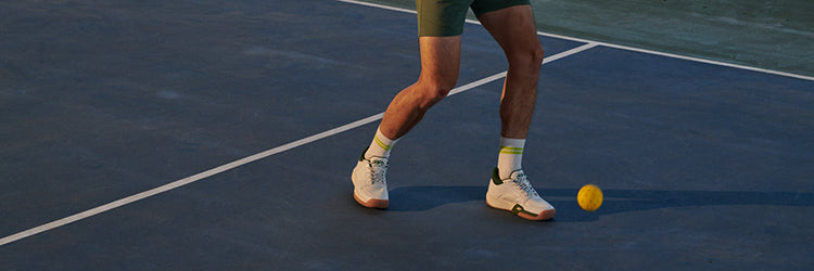 A pickleball player on a blue outdoor court wearing white court shoes, with a yellow pickleball resting near their feet
