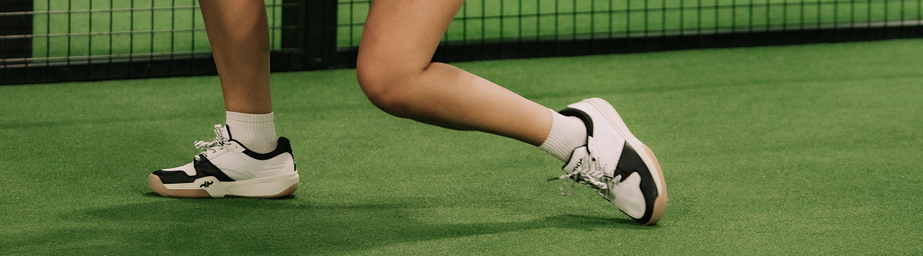 Close-up of a player's feet in white and black court sneakers on a green pickleball court, mid-stride during play