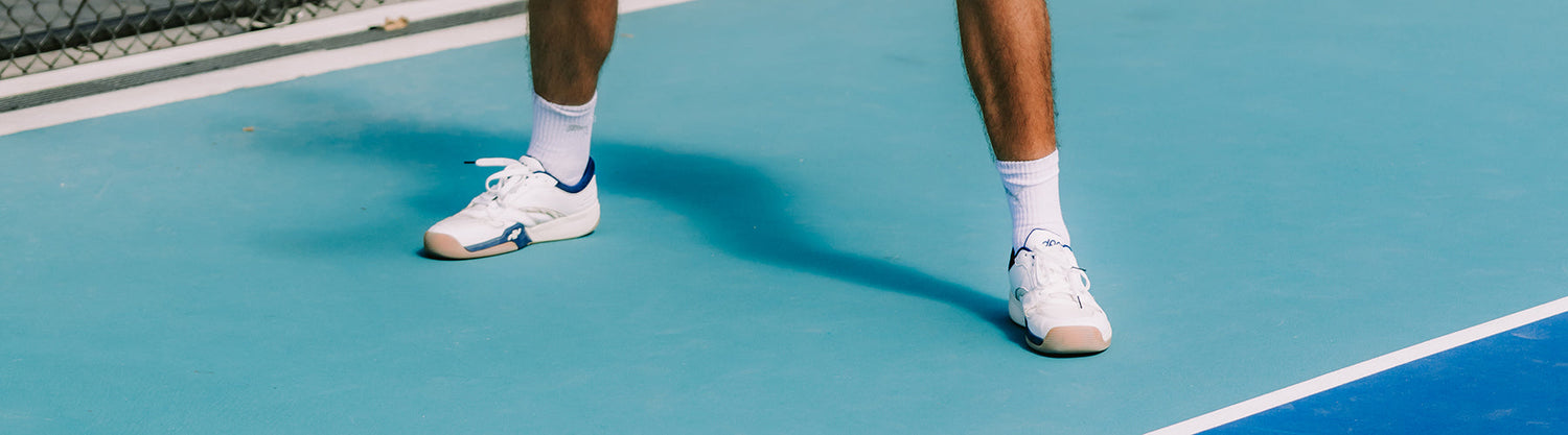 A male pickleball player standing on a blue court wearing white wide-fit sneakers and athletic socks