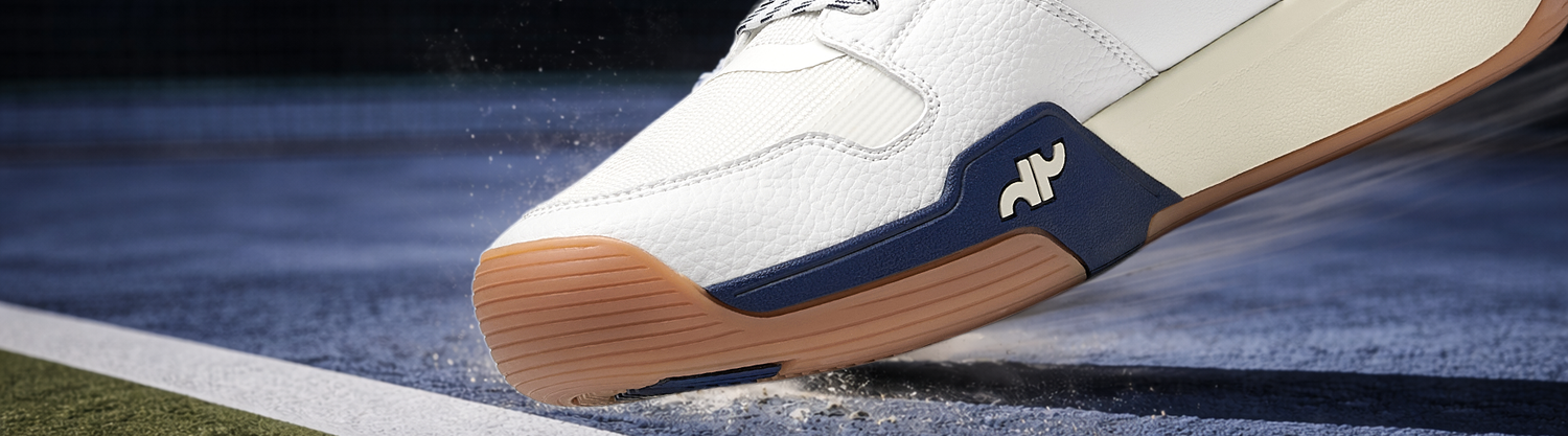 Close-up of a men's white and navy pickleball court shoe mid-slide on a court surface, showcasing grip and stability