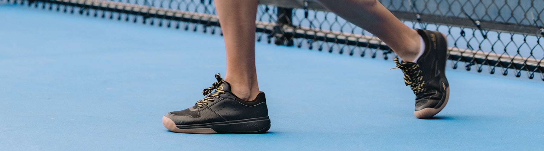 Player wearing DAPS black court shoes with gold laces mid-stride on a blue pickleball court, ideal footwear for grip and agility.