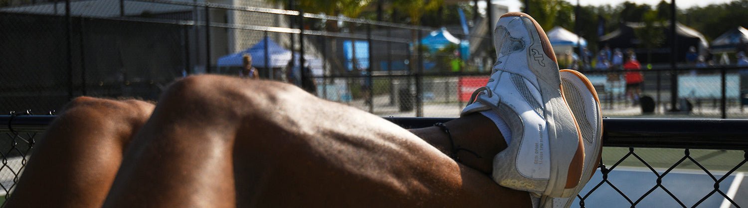 A pickleball player resting courtside, white wide toe box athletic shoes elevated on a chain-link fence with courts and players visible in the background