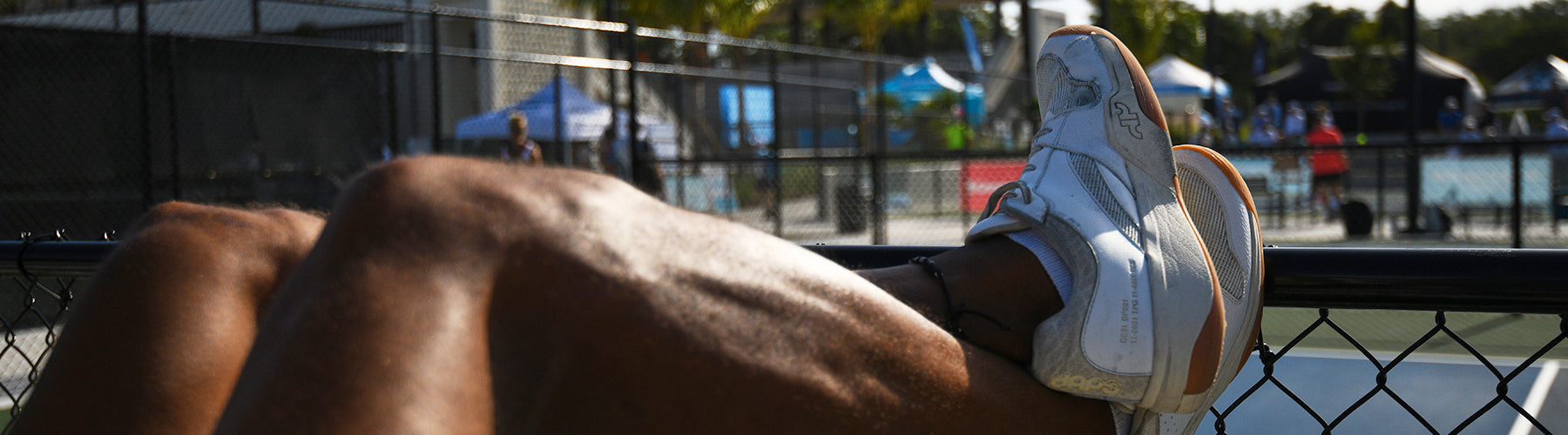 A pickleball player resting courtside, white wide toe box athletic shoes elevated on a chain-link fence with courts and players visible in the background